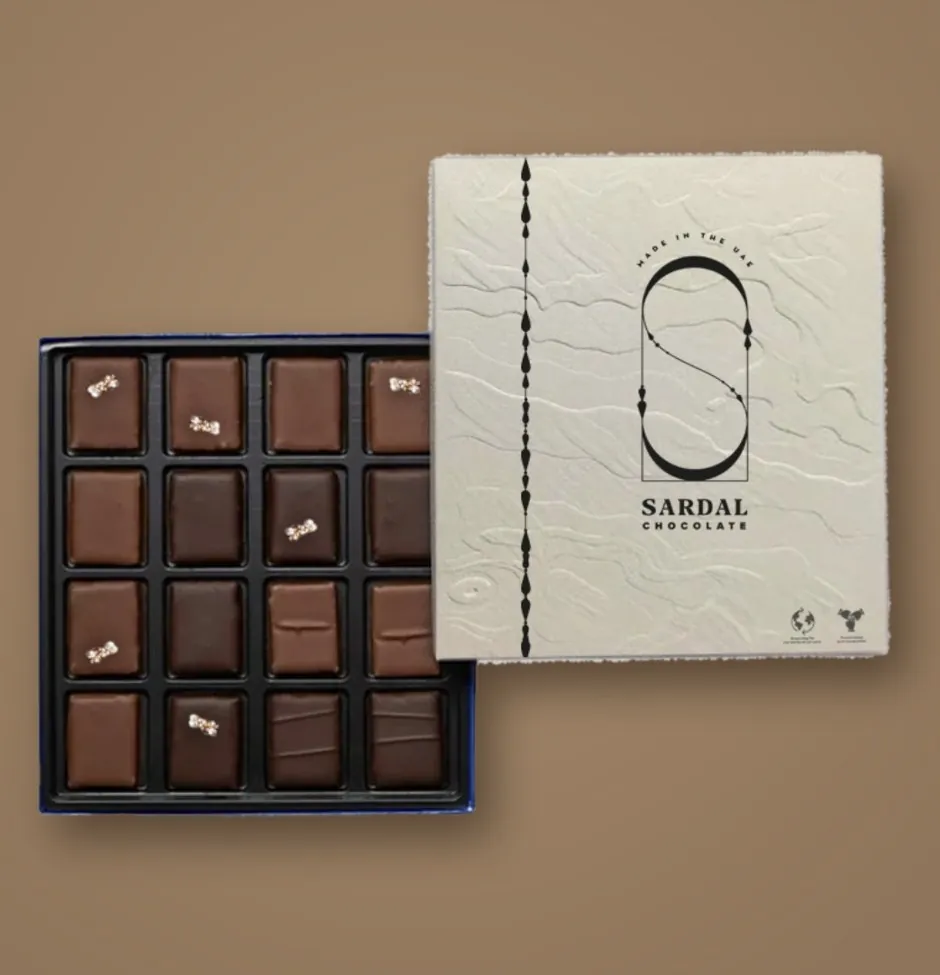 An open box of sixteen square Sardal pralines, some with a pearl-crystal topping, beside its embossed lid